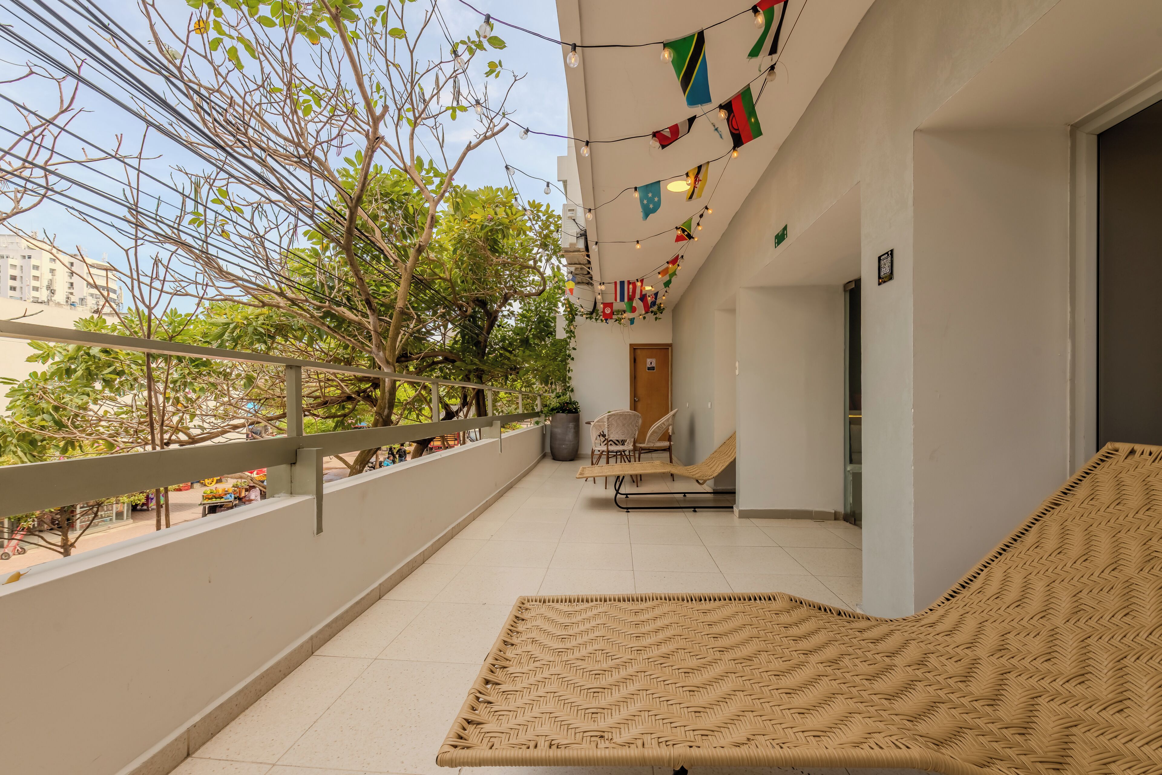 Un balcon avec un canapé et des fauteuils en osier, et une vue sur un arbre et des immeubles. Des drapeaux sont accrochés au-dessus.