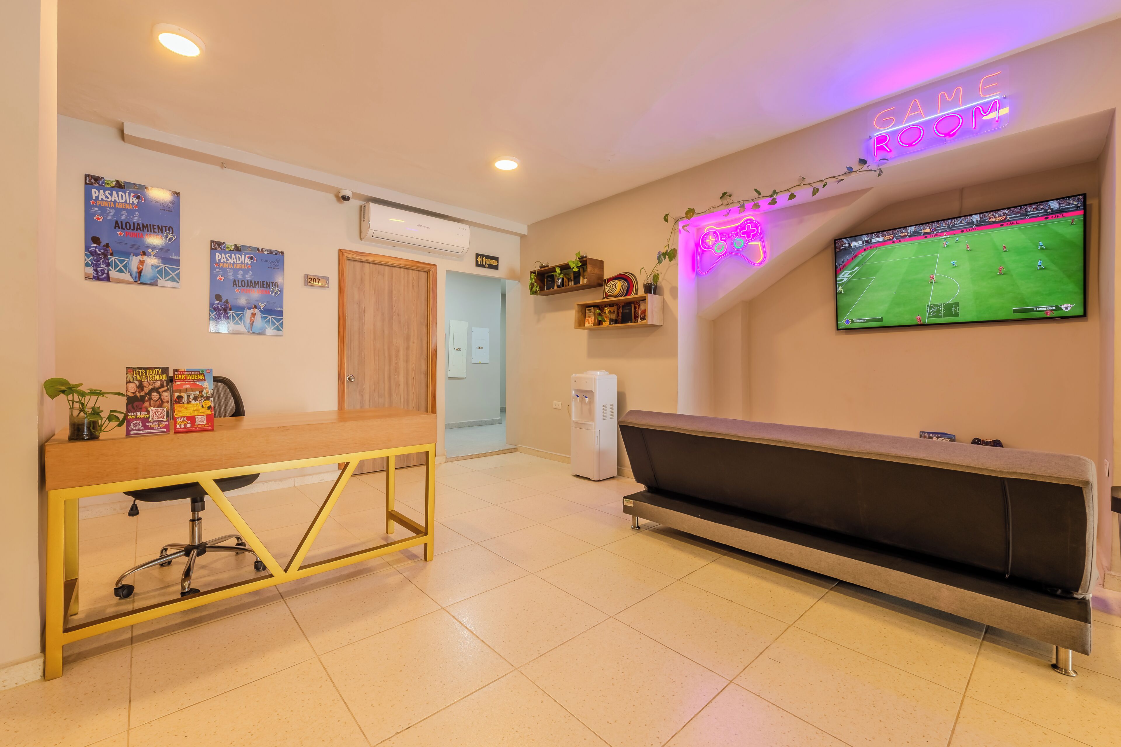 Una habitación con un sofá, un televisor mostrando un evento deportivo, un escritorio de madera con una silla y una estantería en la pared con plantas.