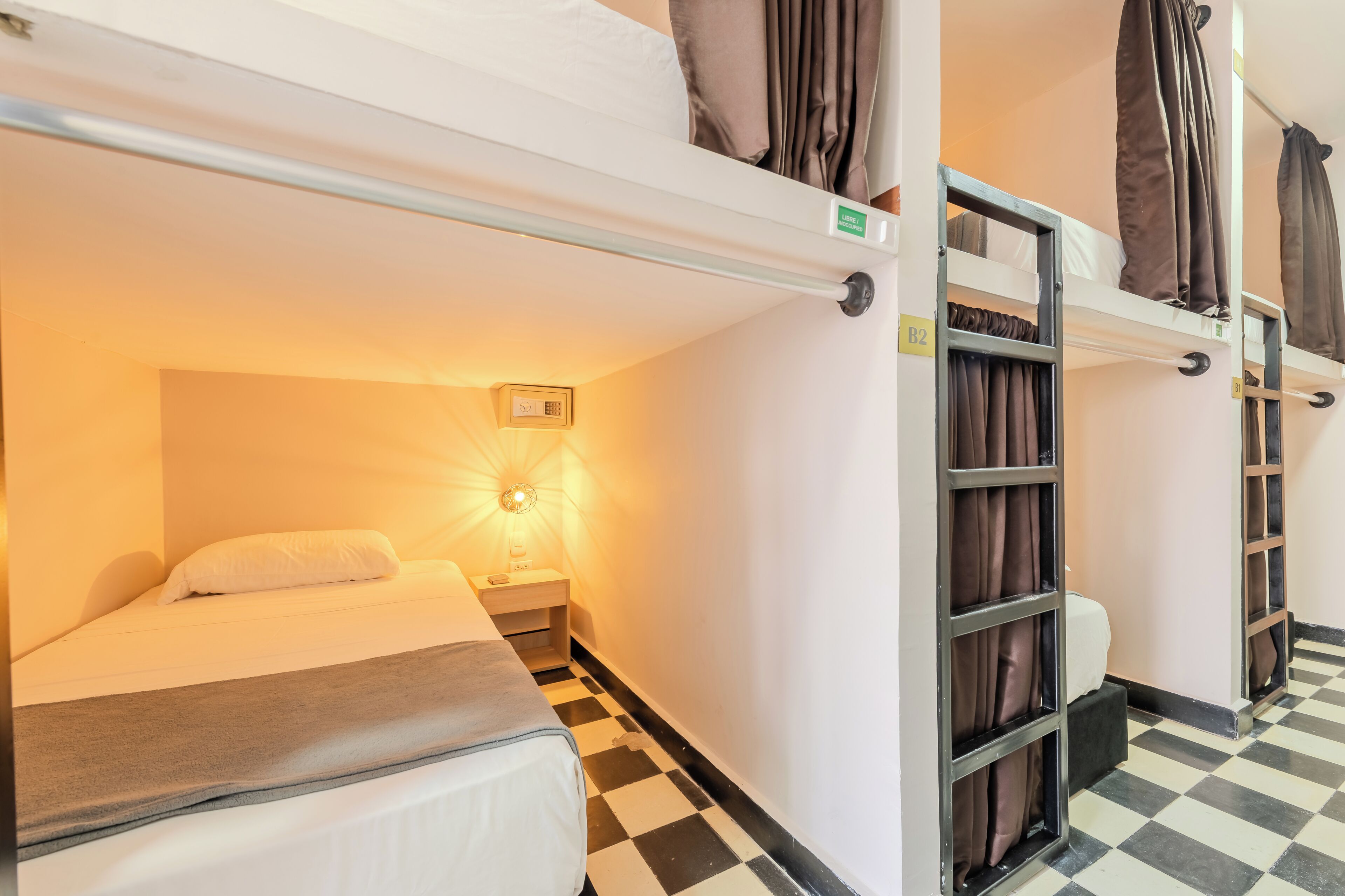A bunk bed room with a checkered floor, a bedside table with a lamp, and a window with curtains.