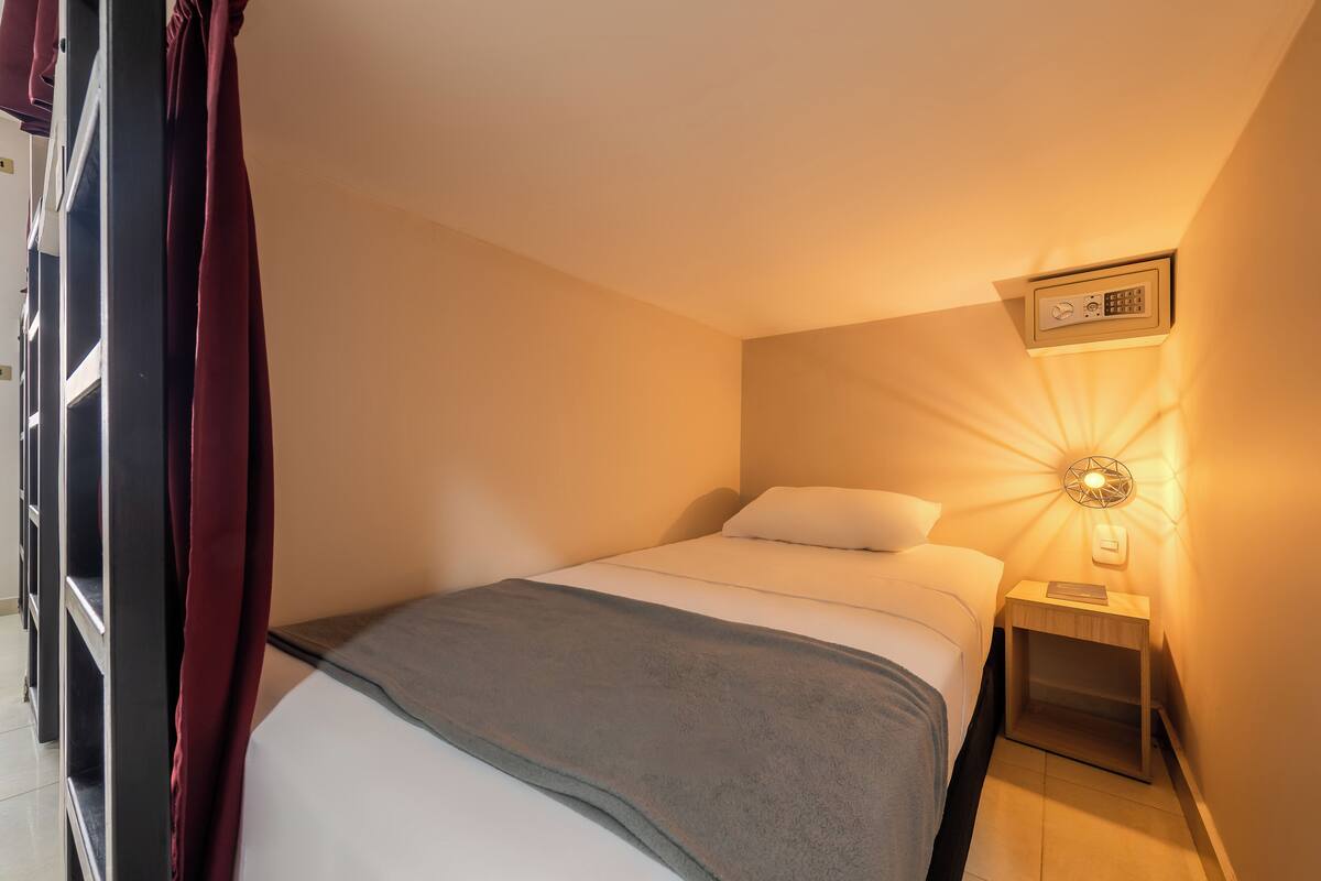 A neatly made bed with a gray blanket, a bedside table with a lamp, and a wall-mounted control panel.