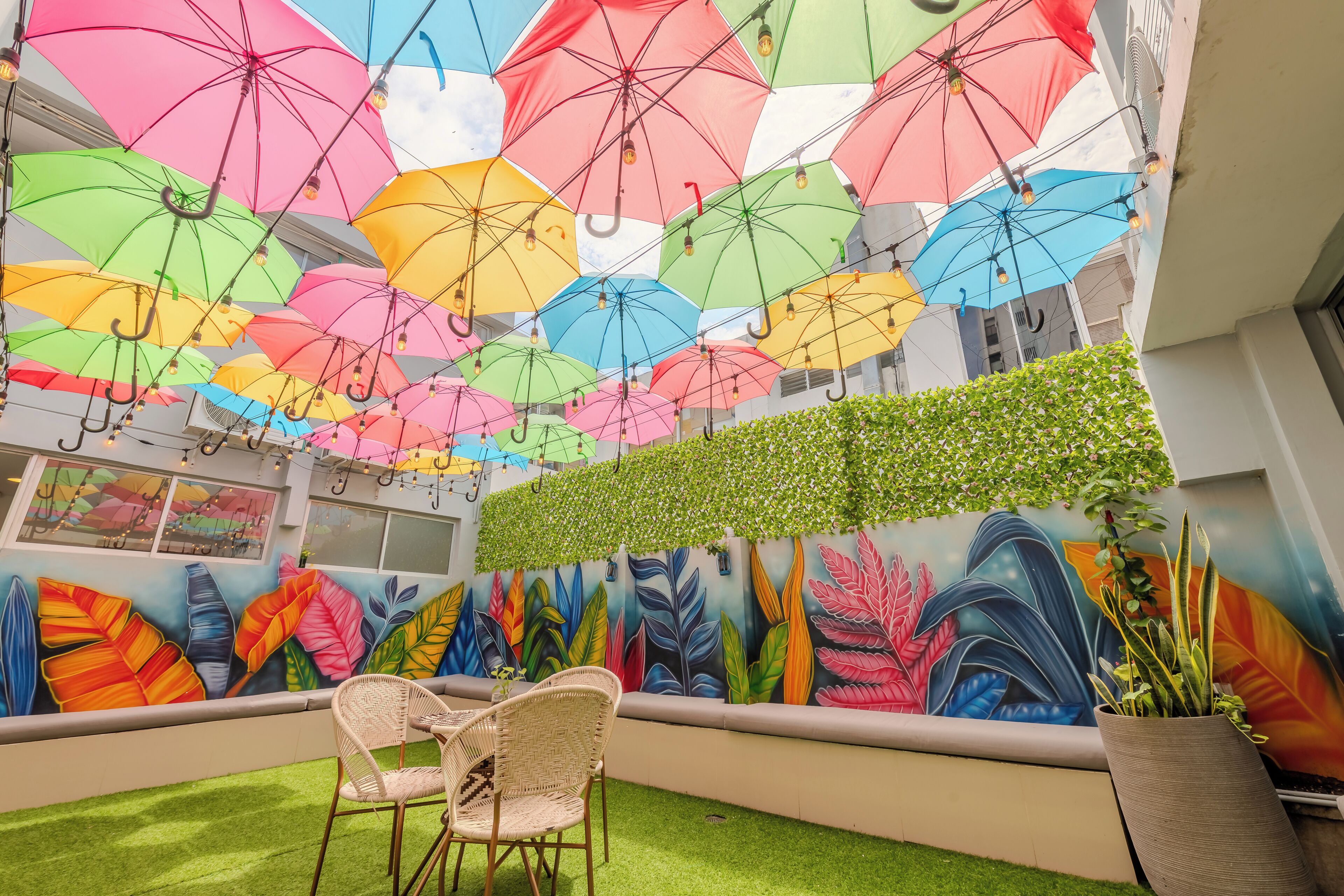 A room with a ceiling adorned with colorful umbrellas, a mural of vibrant leaves, and a seating area with a chair and a table.