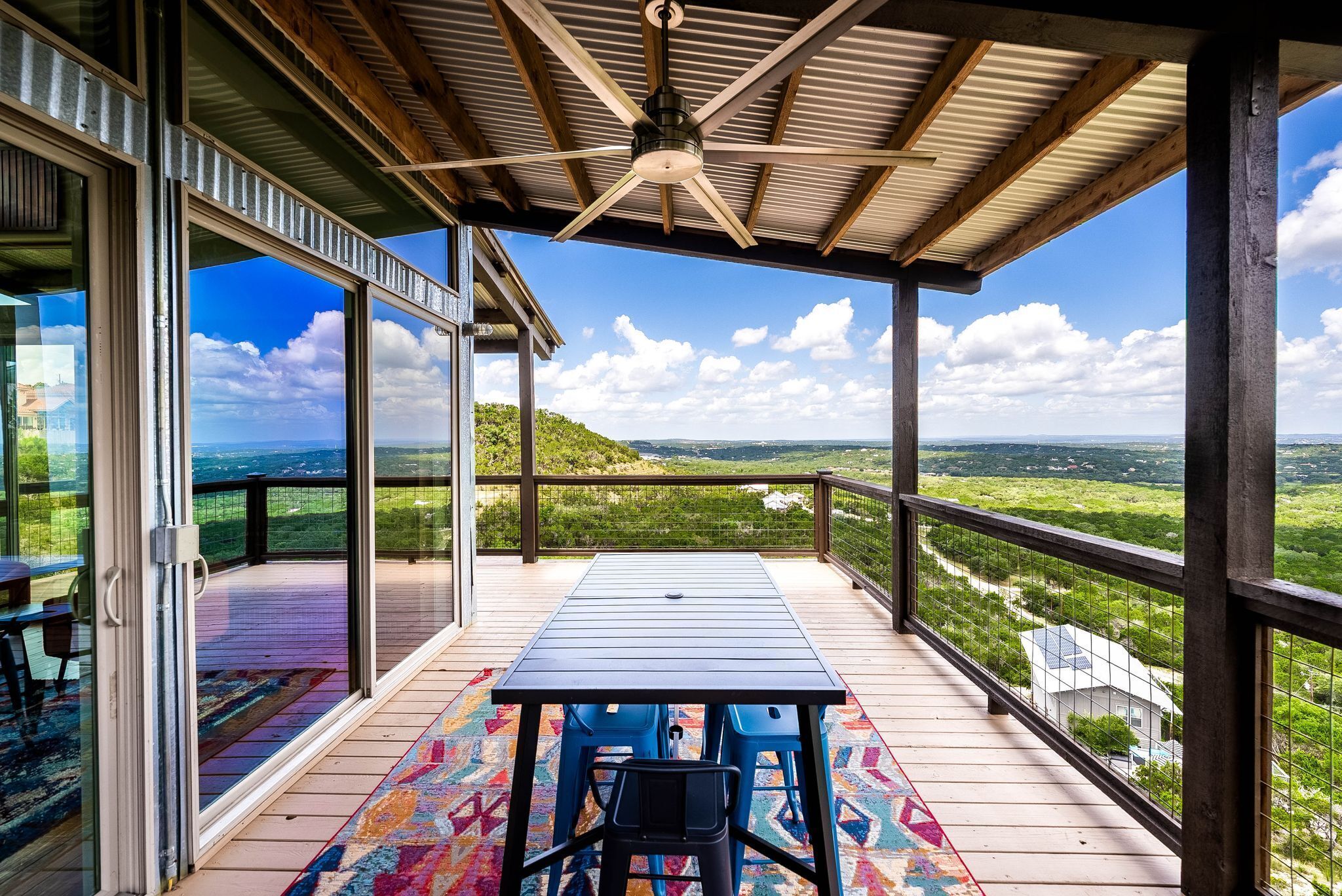 A covered outdoor deck with a table and chairs, a ceiling fan, and a view of a green landscape.