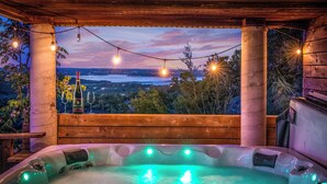 Un bain à remous avec vue sur un lac et des montagnes, entouré d’une terrasse en bois et illuminé de guirlandes lumineuses.
