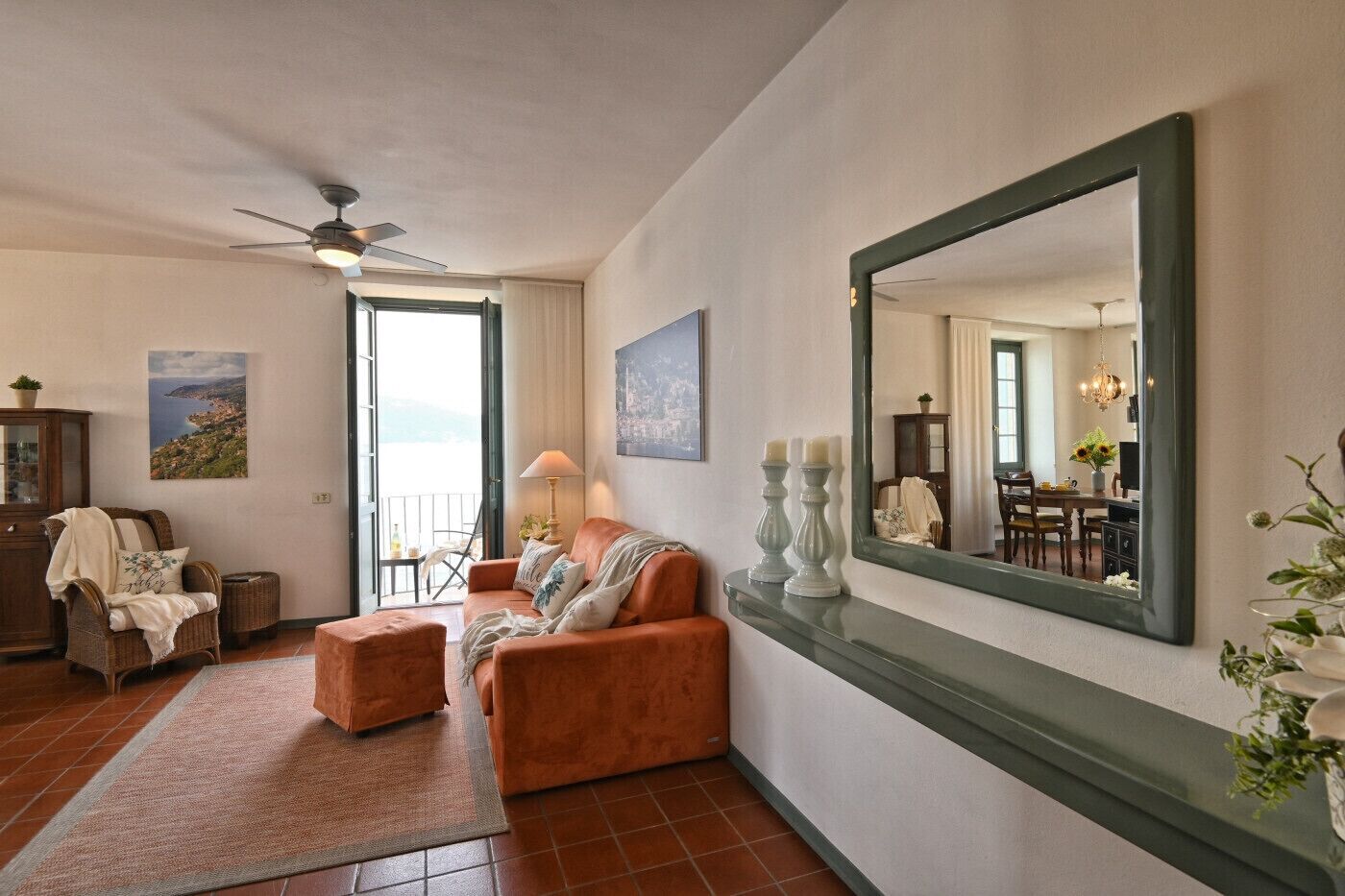 Un salon comprenant un canapé, un fauteuil et un grand miroir. Il y a un balcon avec vue, un ventilateur de plafond et un coin repas visible à travers la fenêtre.
