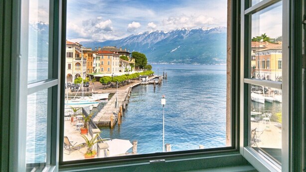 Blick aus dem Fenster auf eine Stadt am Seeufer mit Gebäuden, einem Steg und Bergen im Hintergrund.