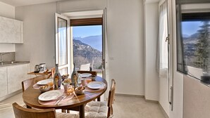 Un comedor con una mesa preparada para cuatro, una cocina con un lavamanos y electrodomésticos, y un balcón con vistas panorámicas a la montaña.