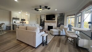 Un salon moderne comprenant un canapé, un pouf et une table basse, une cheminée, un téléviseur et un coin cuisine avec un réfrigérateur et un four à micro-ondes.