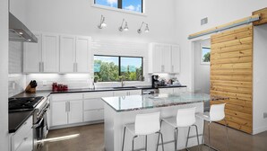 Une cuisine moderne avec des meubles blancs, un grand îlot central doté de tabourets, une cuisinière et une vue sur la verdure par la fenêtre.