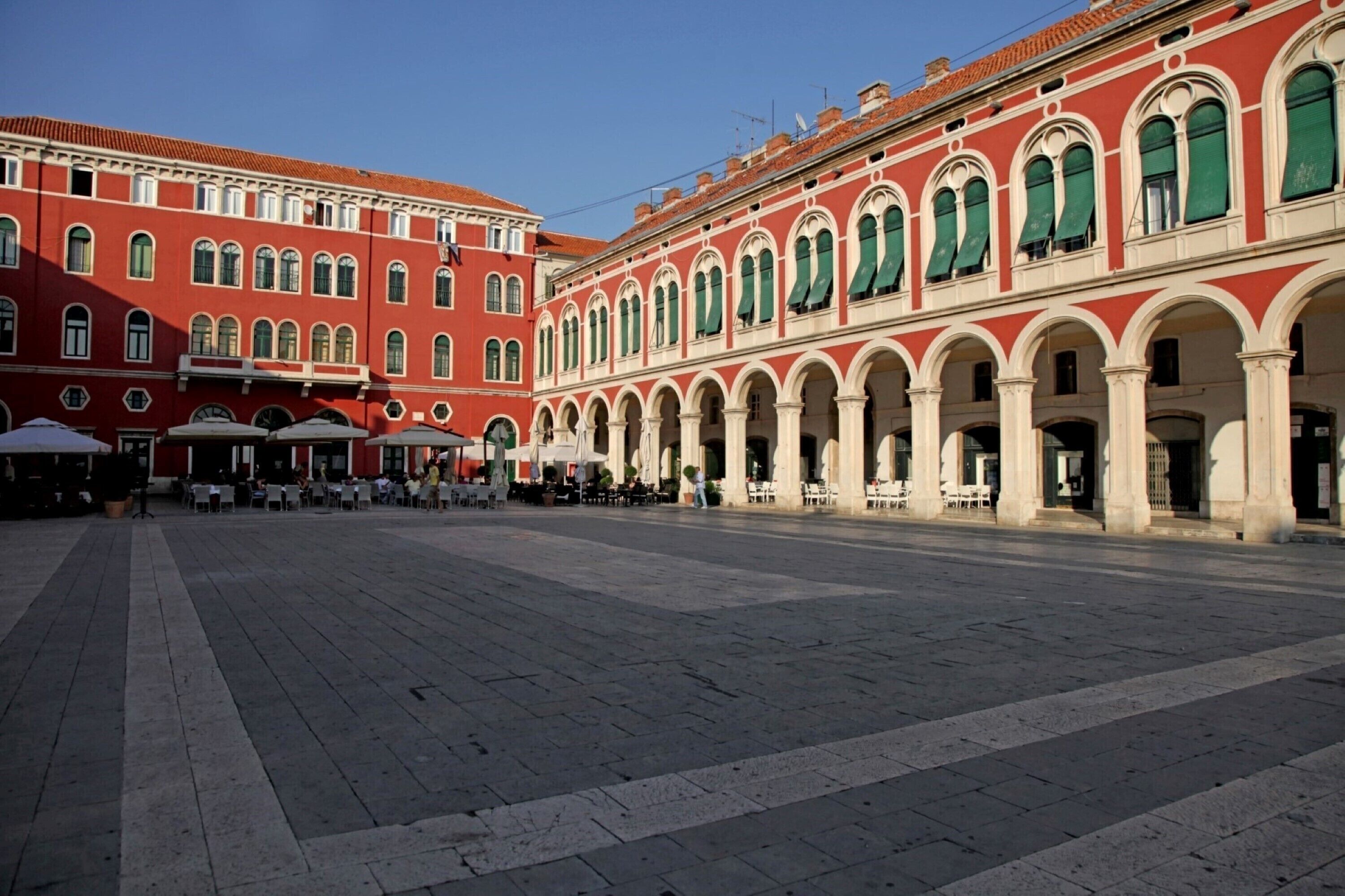A spacious courtyard with red and white buildings, outdoor seating, and a clear sky.
