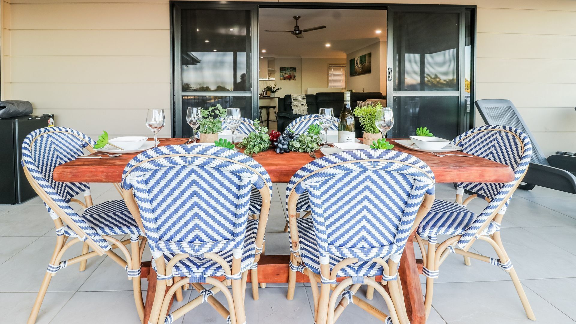 A dining area with a wooden table and blue and white patterned chairs, a glass door leading to a kitchen, and a ceiling fan.