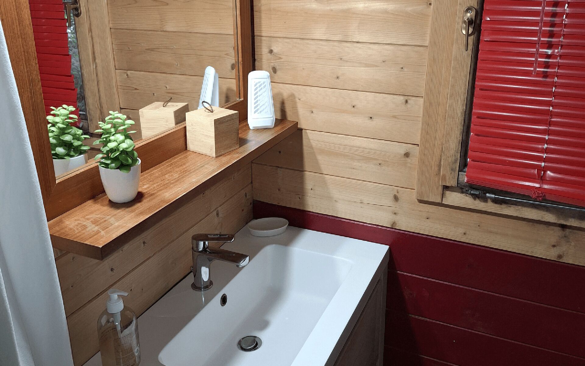 A bathroom with a wooden vanity, a white sink, a mirror, a toilet, and a window with red blinds.