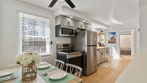 Una cocina moderna con electrodomésticos de acero inoxidable, un comedor con una mesa lista para dos personas, y vistas al exterior a través de una ventana con persianas.