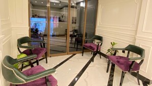 Un hall d’hôtel avec un sol en marbre, une entrée avec une porte en verre et deux fauteuils verts dotés de coussins violets.