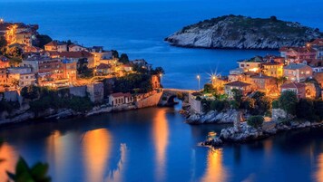 Un pueblo costero al atardecer, con edificios a lo largo del paseo marítimo, un puente y una pequeña isla al fondo.