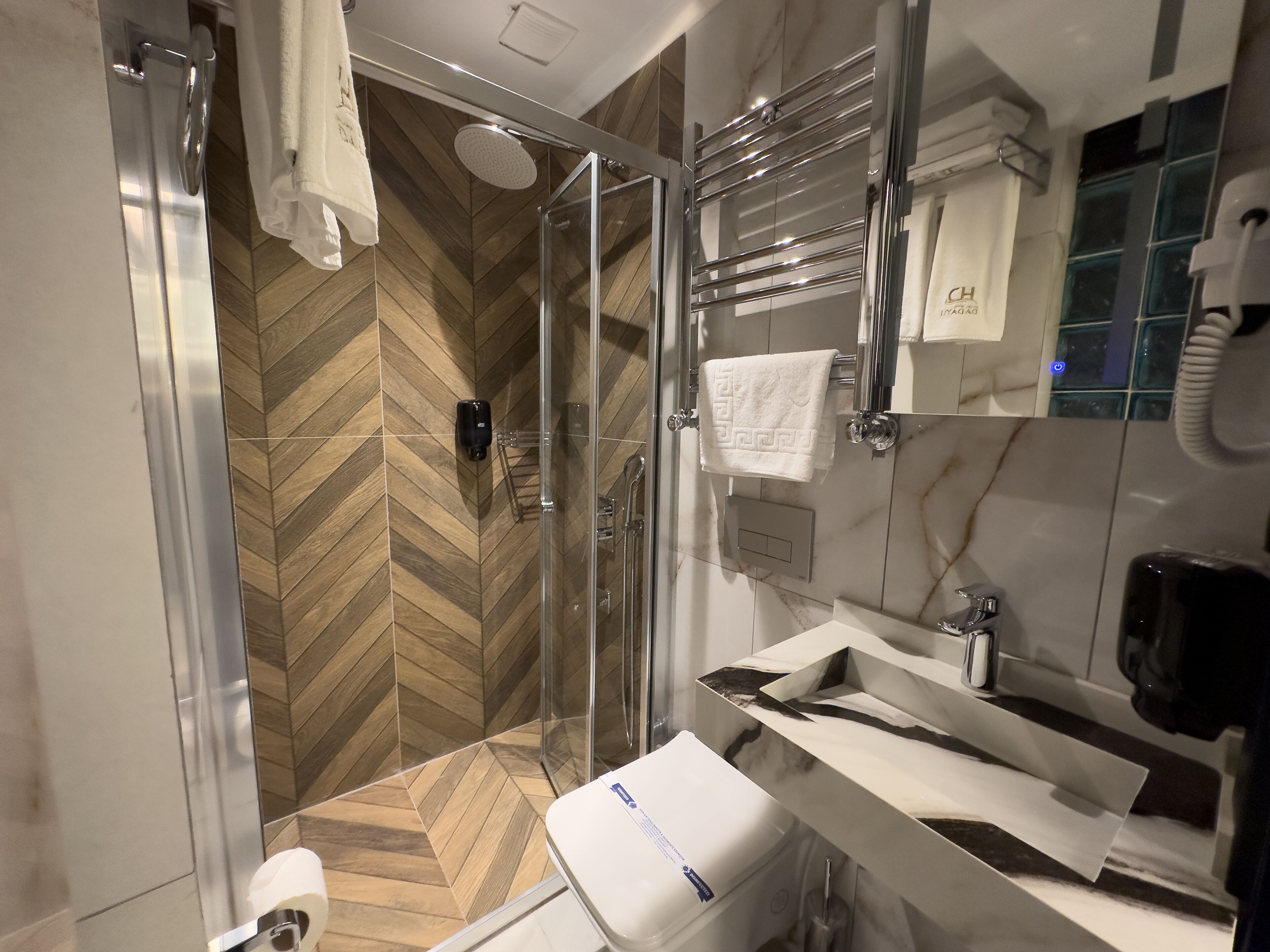 Un baño moderno con piso de madera en patrón chevron, cabina de ducha de vidrio, lavamanos blanco con encimera de mármol y toallero con una toalla blanca.