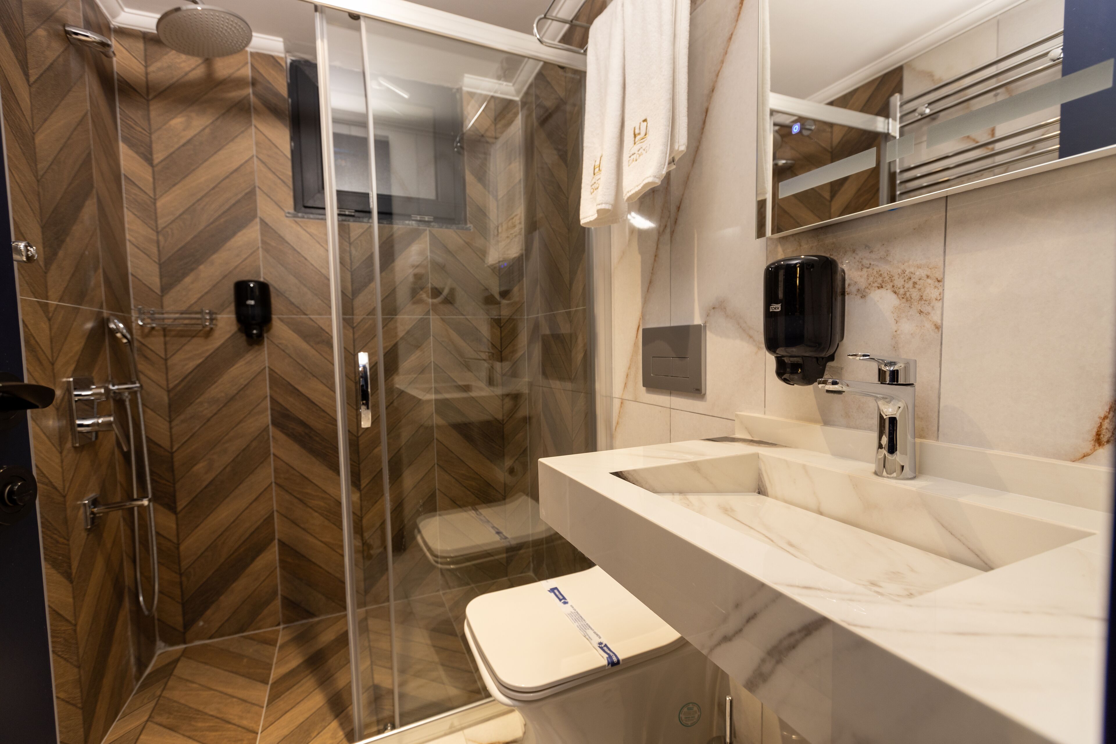 A modern bathroom with a herringbone wood floor, a glass shower enclosure, a white toilet, a marble sink, and a hand dryer.