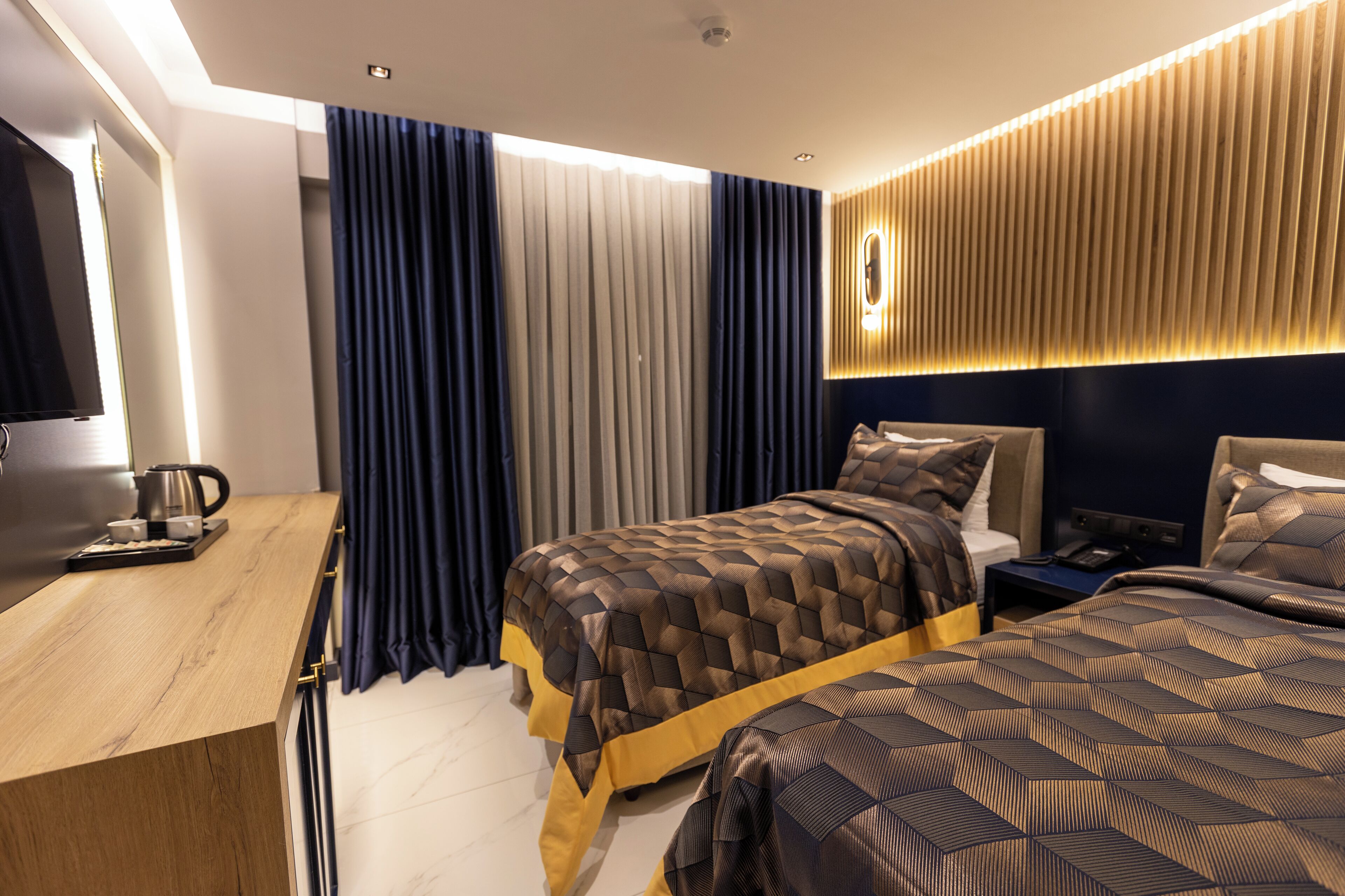 A modern hotel room with a wooden desk, a bed with patterned bedding, a nightstand, and a wall-mounted light fixture.