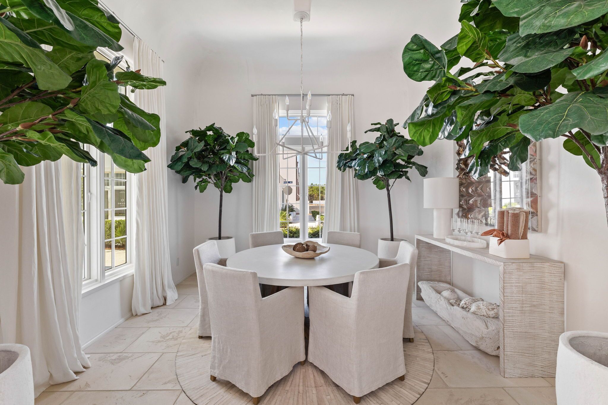 A dining area with a round table, white chairs, and large potted plants.