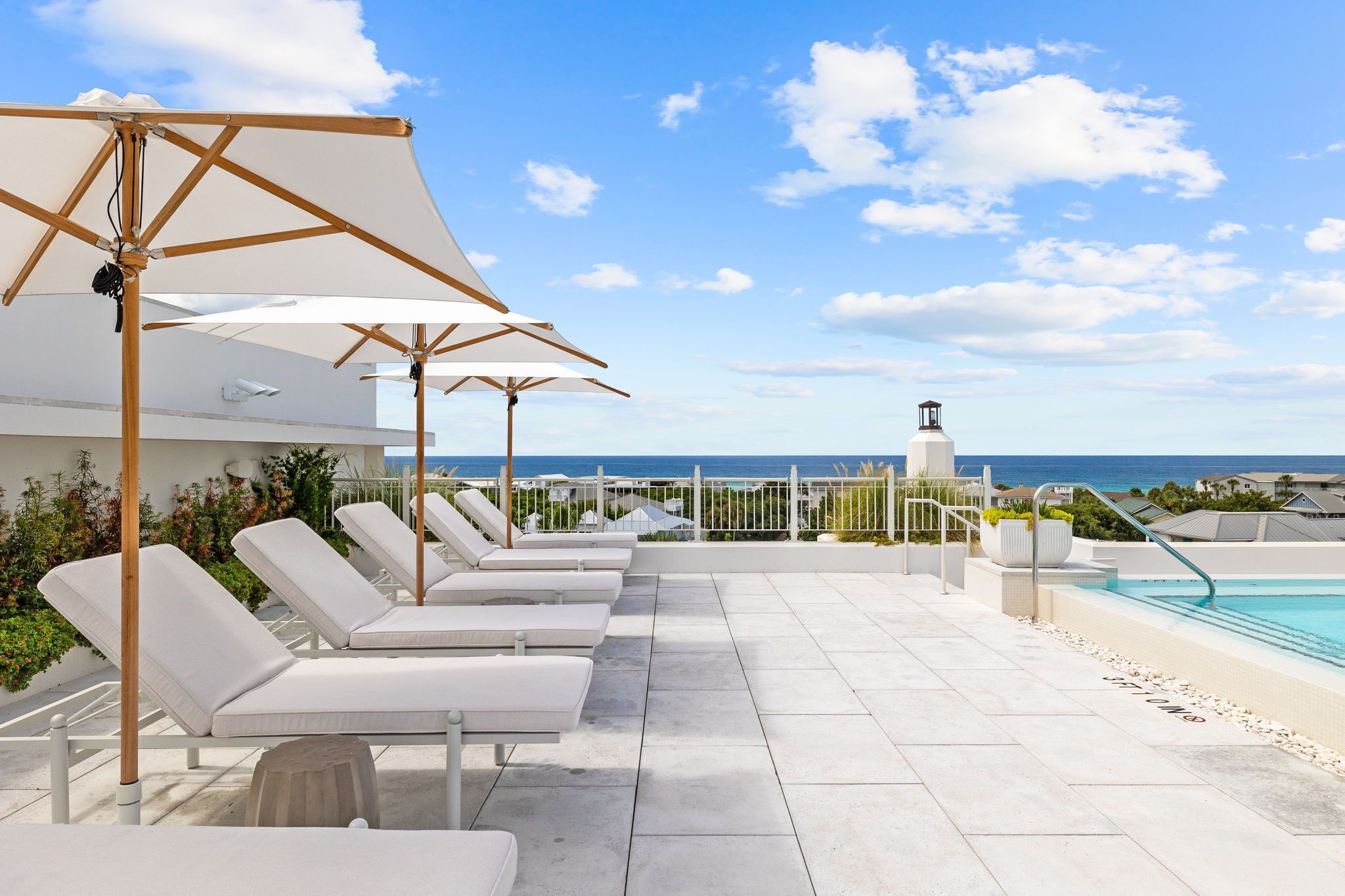 Un espace aménagé au bord de la piscine, avec des chaises longues et des parasols, une vue dégagée sur la mer et un bâtiment doté d’un clocher.