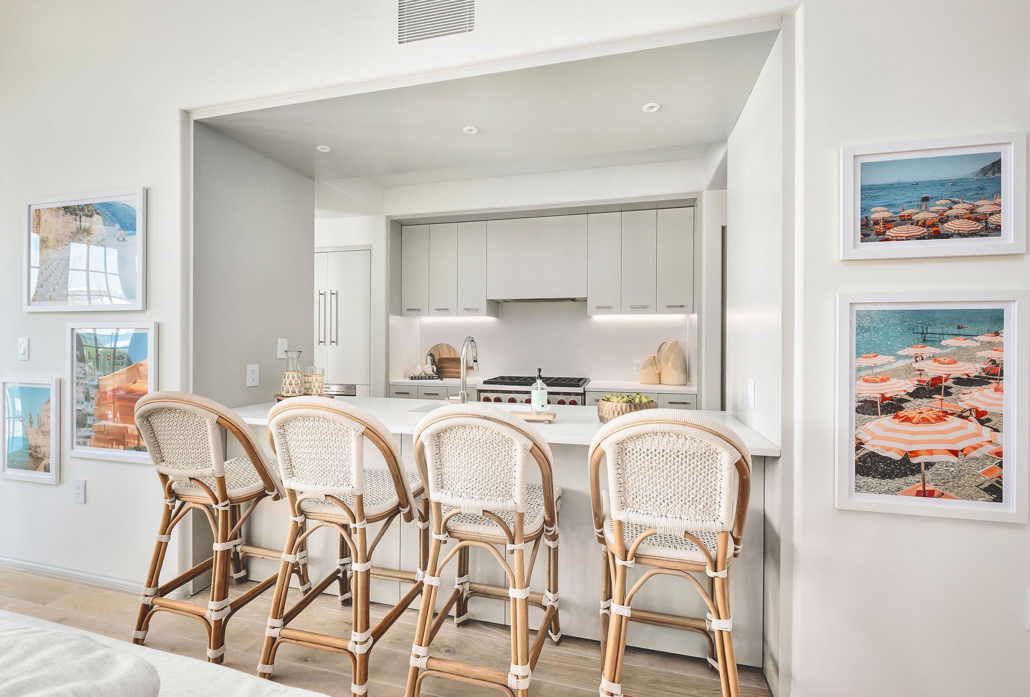 A modern kitchen with white cabinets, a central island with bar stools, and framed coastal artwork on the walls.