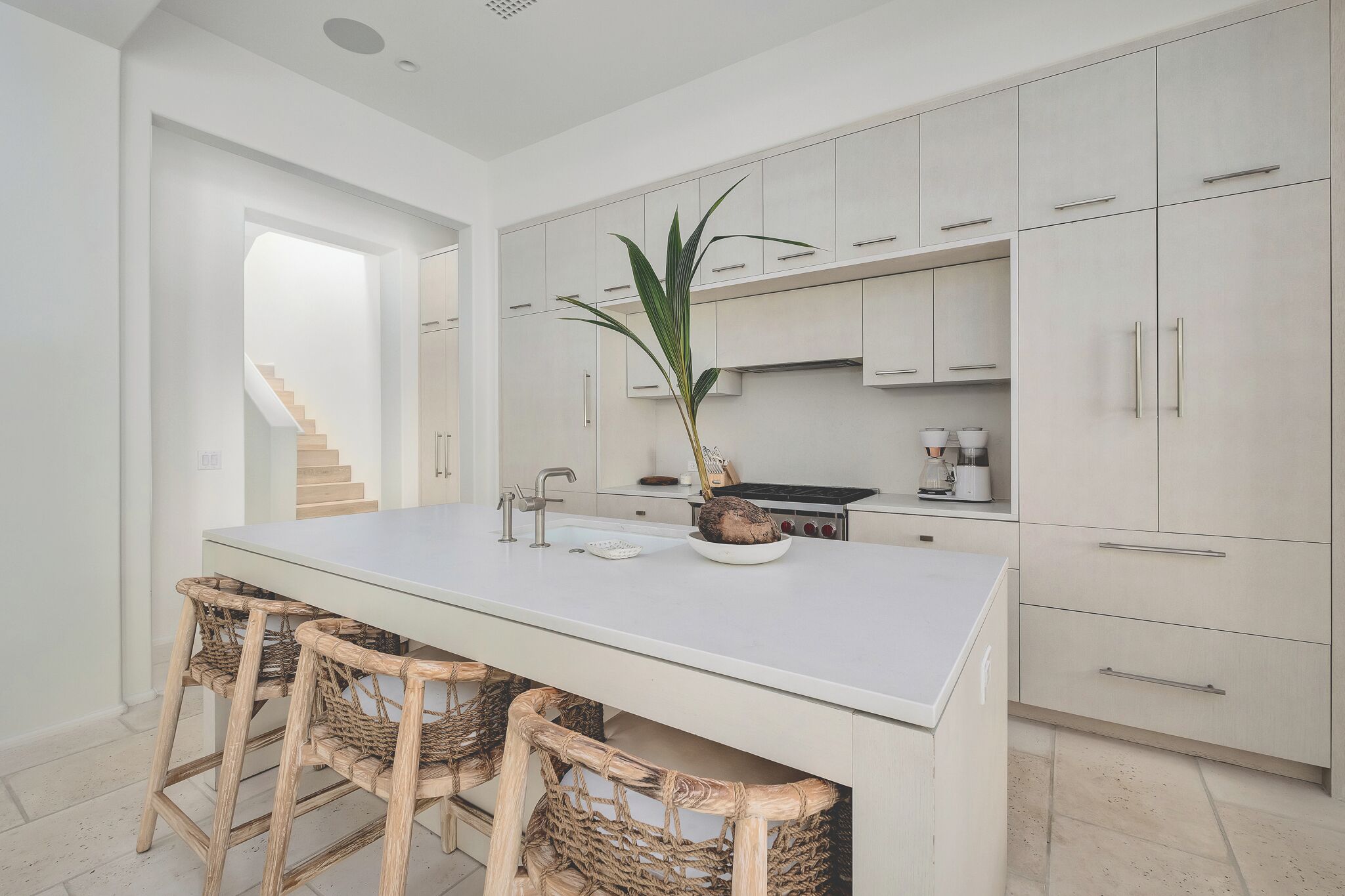 Una cocina moderna con una isla central, sillas de mimbre y una escalera que sube a otro nivel.