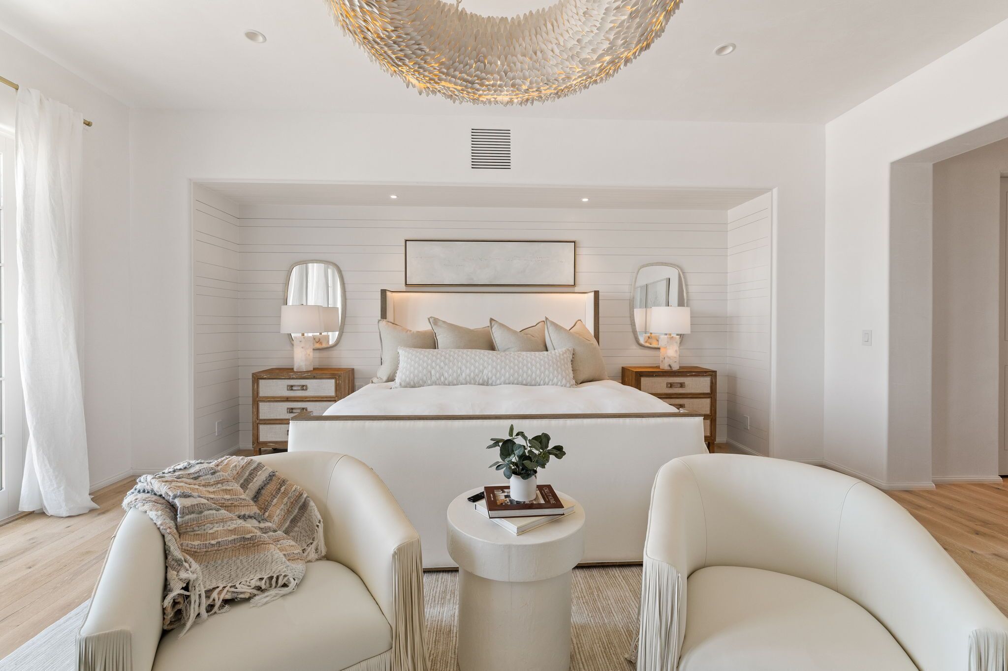 A modern bedroom with a large bed, bedside lamps, a small round table with a book and plant, and two armchairs.