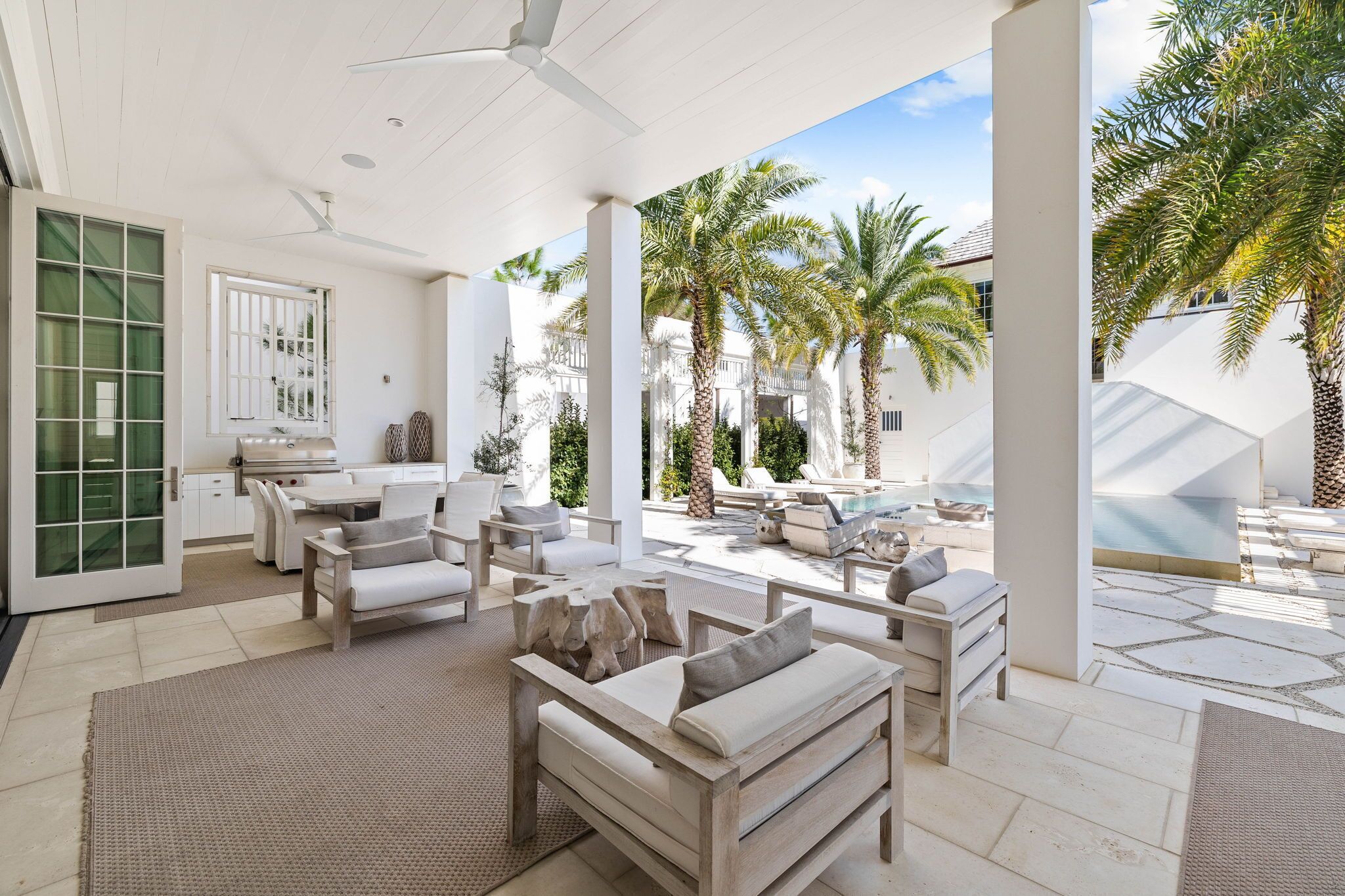 A covered outdoor seating area with a pool, palm trees, and a modern kitchen visible through the glass doors.
