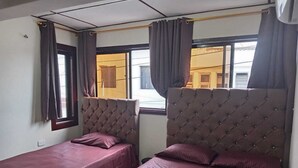A room with two beds, each with a tufted headboard and covered with a maroon bedspread. There are curtains on the windows, and a visible window reveals a building outside. - La Perla del Caribe  (Riohacha)