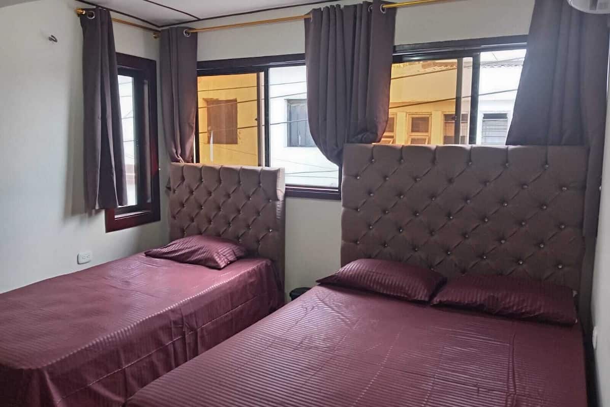 Une chambre avec deux lits, chacun doté d’une tête de lit capitonnée et recouvert d’une couvre-lit de couleur bordeaux. Des rideaux sont accrochés aux fenêtres, et une fenêtre donne sur un bâtiment situé à l’extérieur.