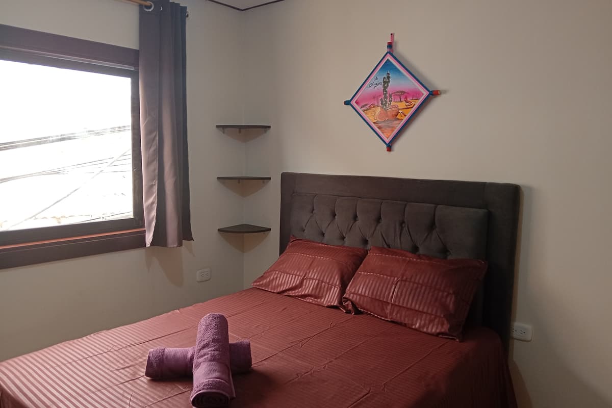 Une chambre à coucher avec un lit, une fenêtre avec des rideaux, une étagère fixée au mur et un objet décoratif.