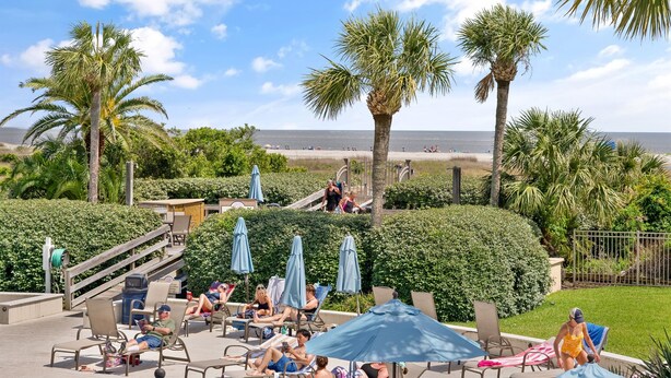 A pool area with lounge chairs and umbrellas, palm trees, and a view of the beach. - Fall Special | Condo with pool, steps to the beach (St Simons Island)