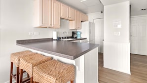 A modern kitchen with a granite countertop, wooden stools, and light wooden cabinets. - Fall Special | Condo with pool, steps to the beach (St Simons Island)