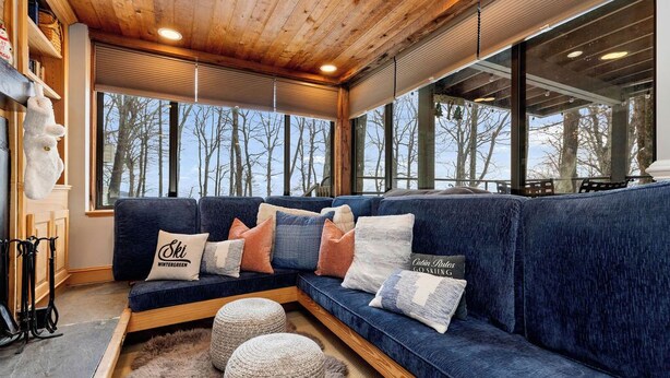 A cozy living room with a blue sectional sofa, wooden beams, large windows, and a view of trees.