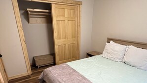 A bedroom with a wooden floor, a bed with white bedding, a wooden wardrobe, and a small bedside table.