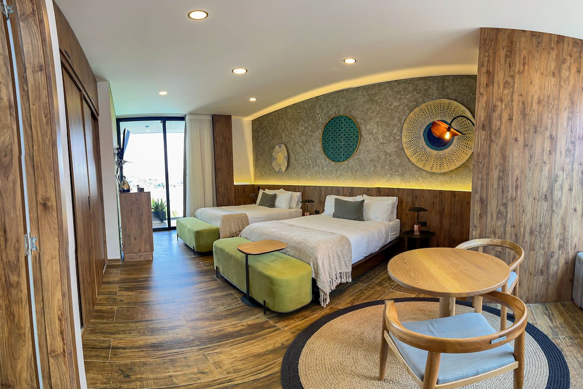 A modern hotel room with a large bed, two smaller beds, a wooden floor, and a round wooden table with chairs.