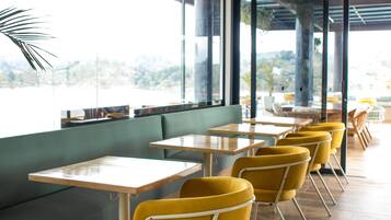 Interior moderno de restaurante con mesas de madera y sillas amarillas, ventanas amplias y plantas colgantes.