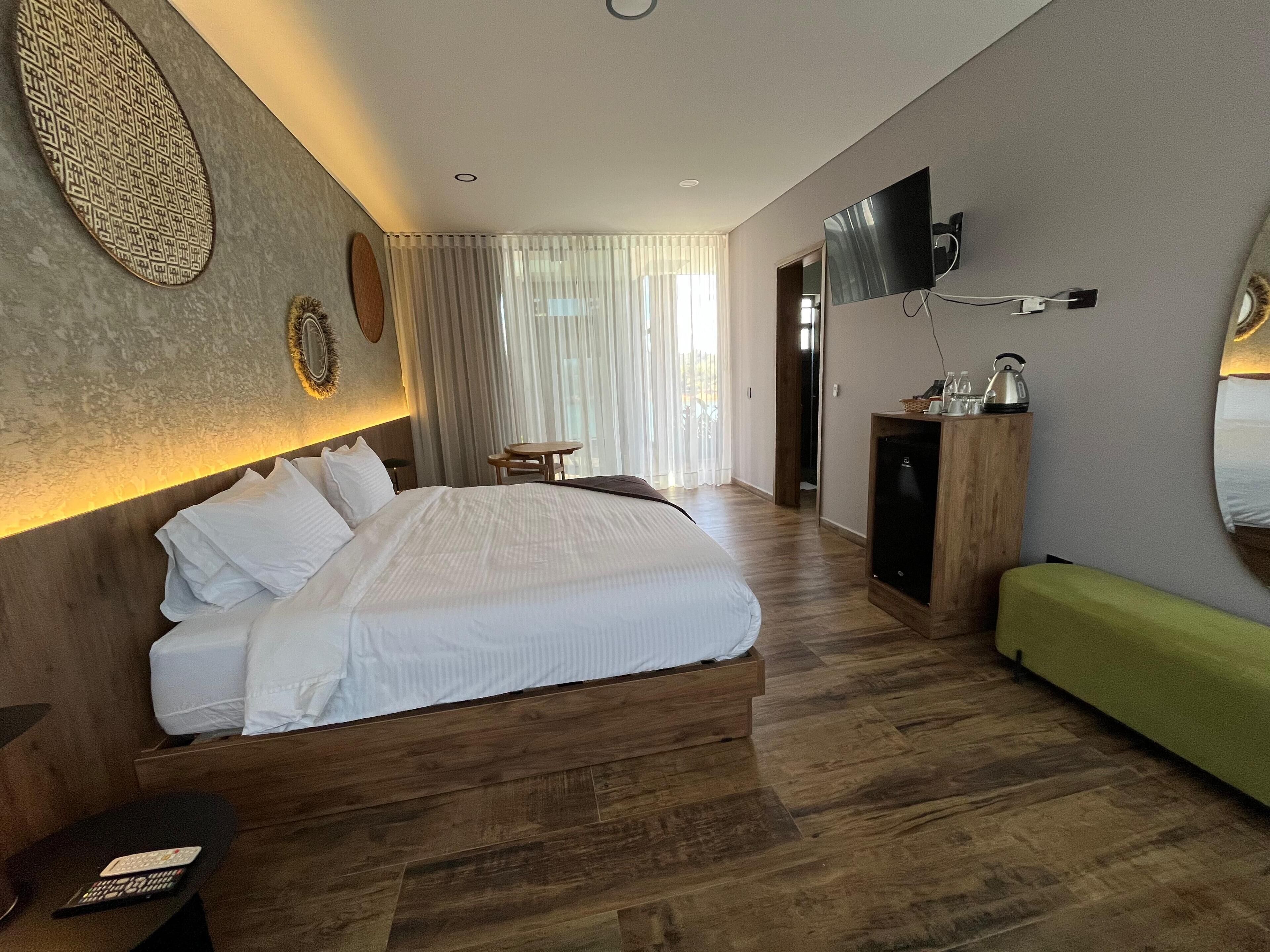 A hotel room with a large bed, a wooden headboard, a flat-screen TV mounted on the wall, a small wooden table, and a green bench.