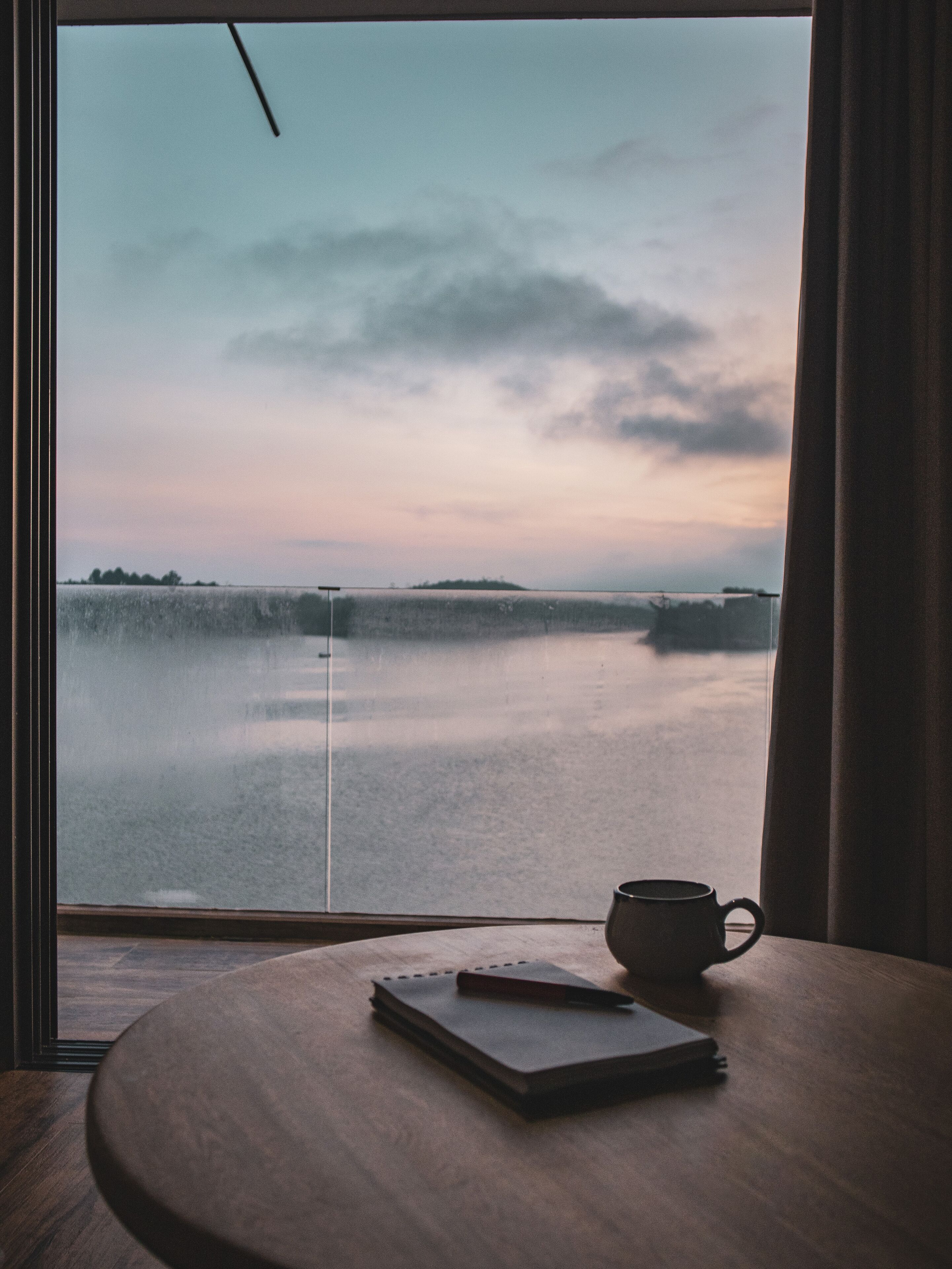 A room with a large window overlooking a body of water, a wooden table with a notebook and a mug, and a curtain partially drawn to the side.