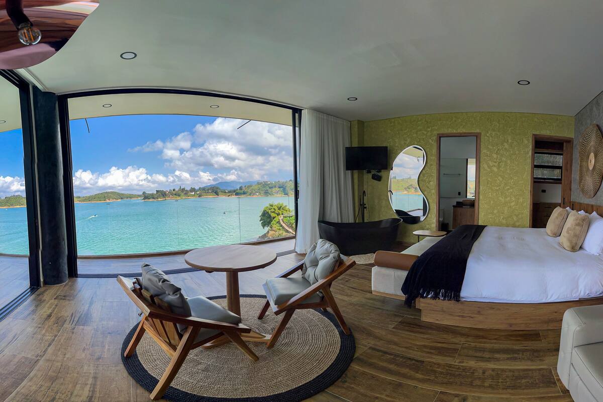 A bedroom with a large window, a bed, a TV, and a seating area with a view of the sea.