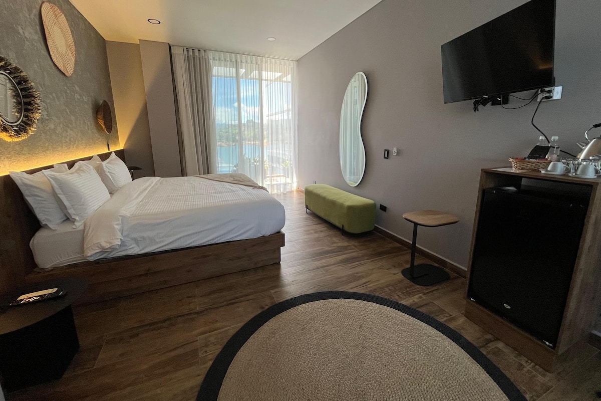 A modern hotel room with a large bed, a desk with a flat-screen TV, a small seating area, and a view of the outdoors.