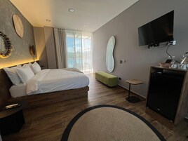 A modern hotel room with a large bed, a desk with a flat-screen TV, a small seating area, and a view of the outdoors.