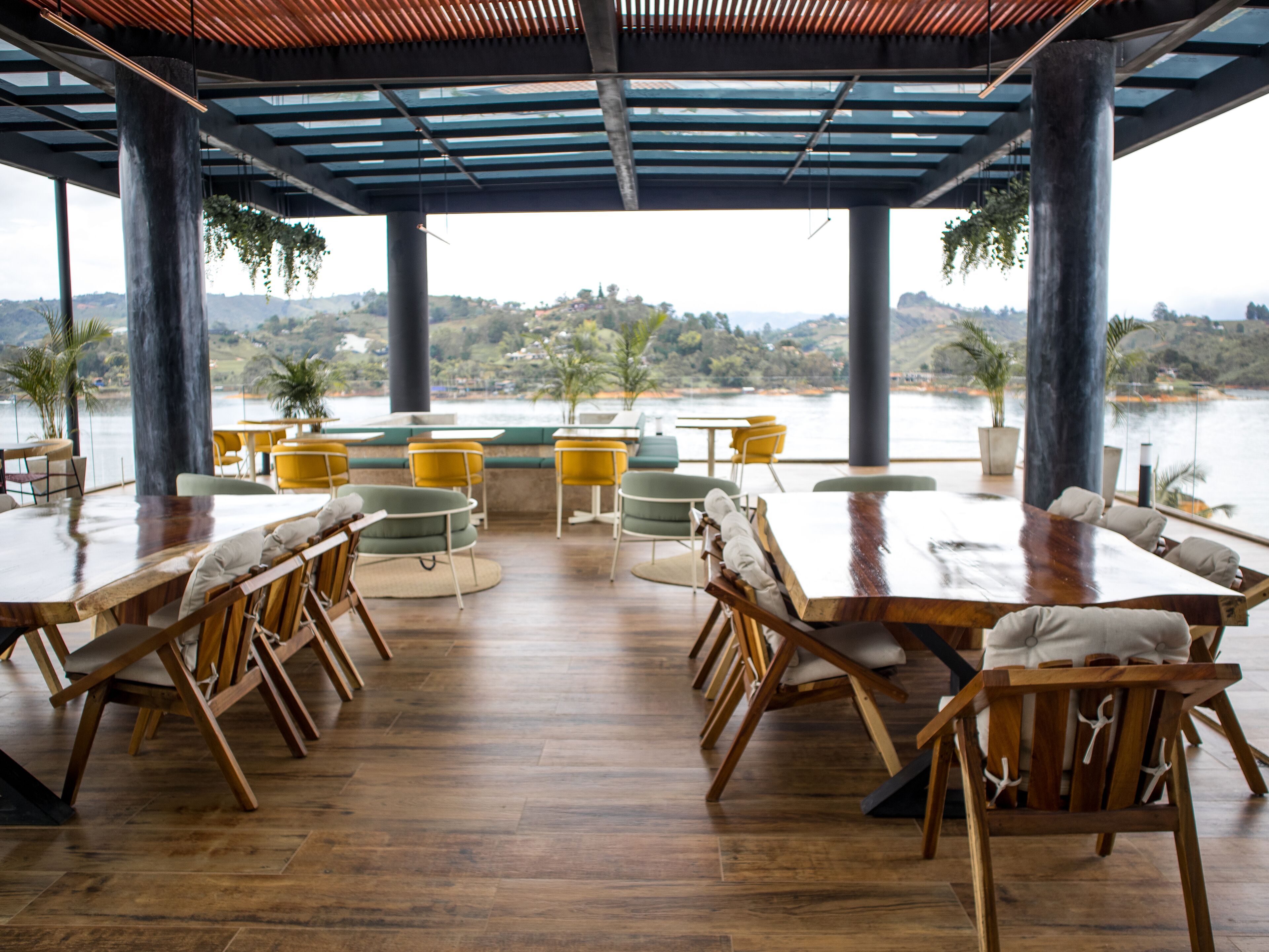 A spacious outdoor dining area with wooden tables and chairs, surrounded by large pillars, overlooking a scenic view of hills and a body of water.