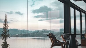 A modern room with a large window overlooking a lake and hills. There is a wooden chair and a small table.