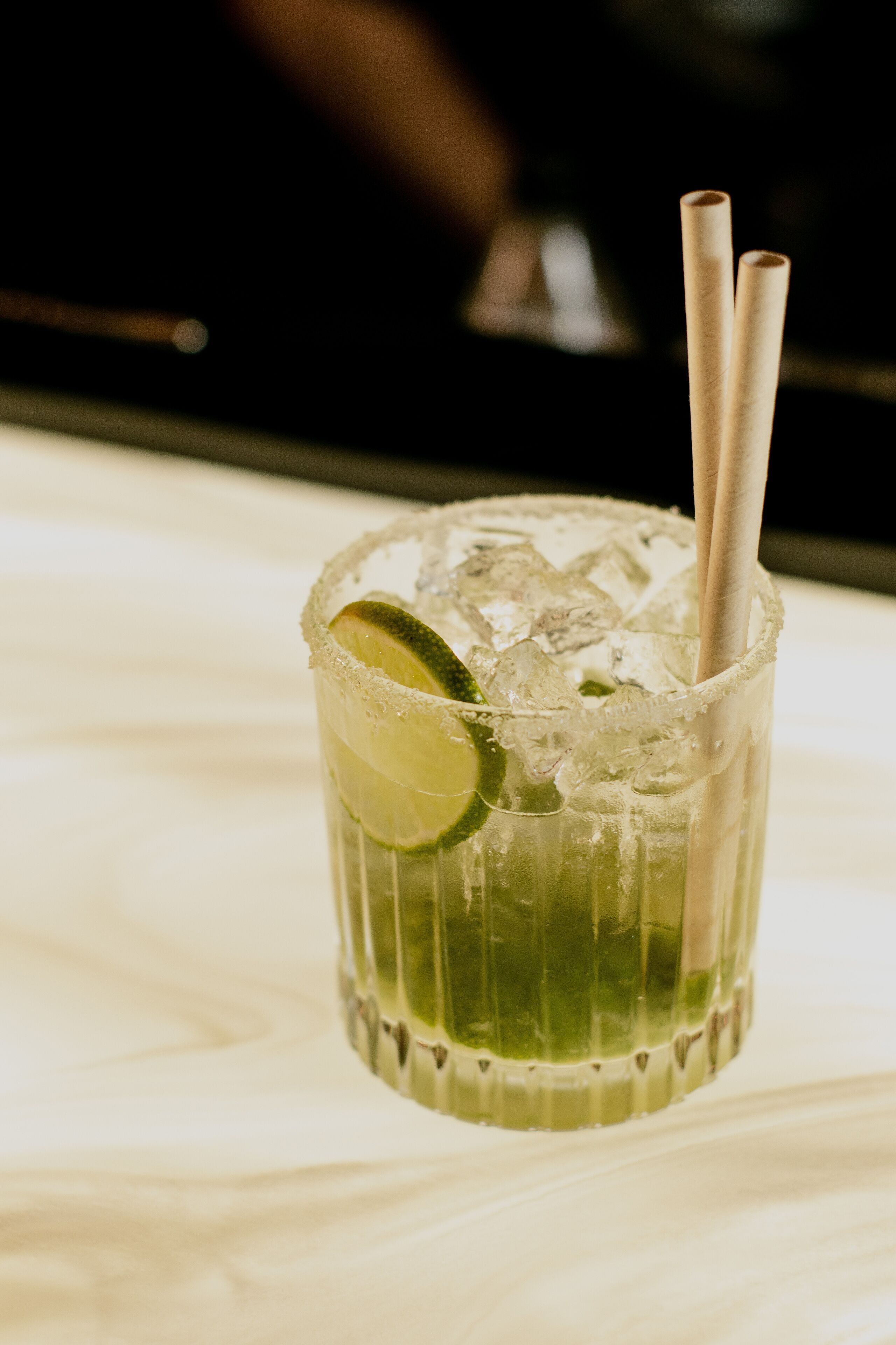 A glass of a green cocktail with lime, ice, and two straws on a table.