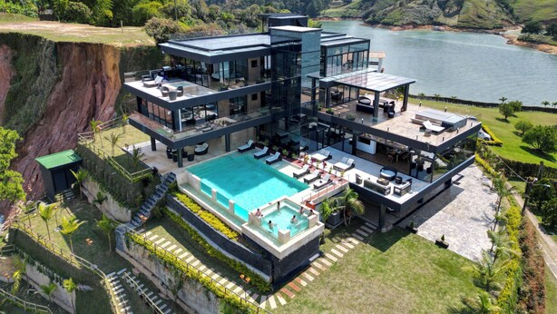 A modern multi-story house with a large swimming pool, outdoor seating, and a view of a lake and mountains.