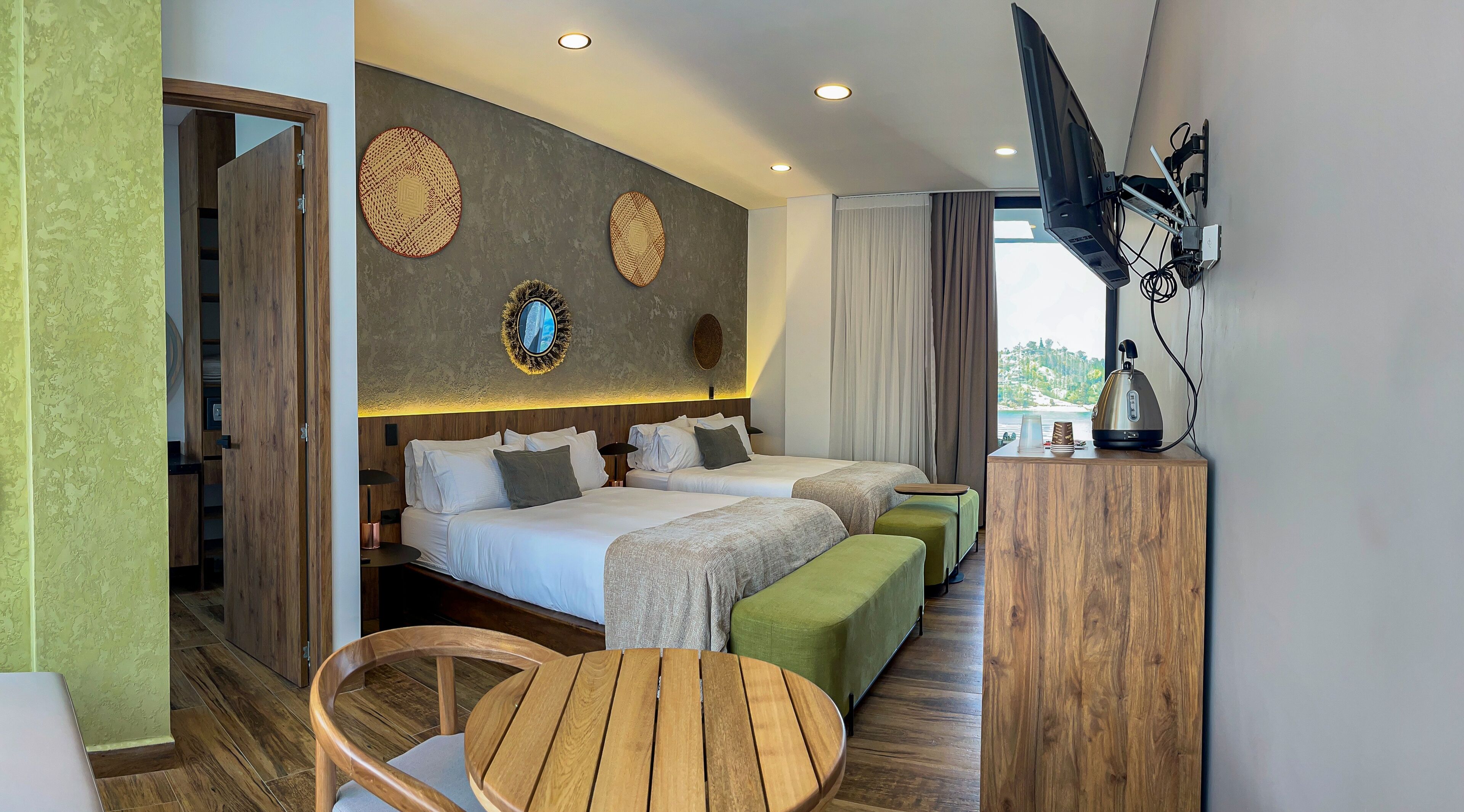 A hotel room with two beds, a wooden table, a green ottoman, and a view of the sea.