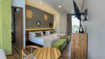 A hotel room with two beds, a wooden table, a green ottoman, and a view of the sea.