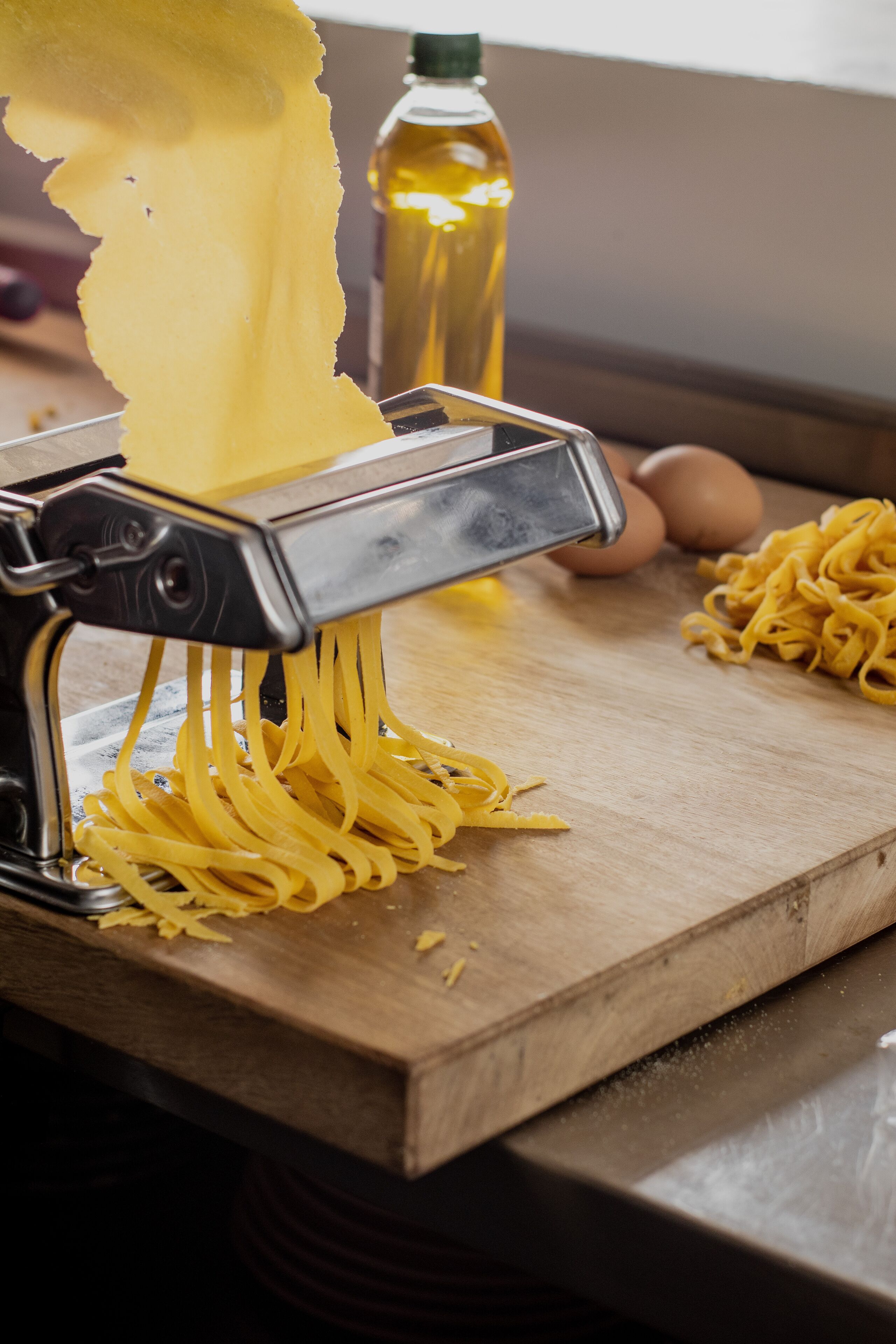 A kitchen with a pasta machine on a wooden countertop, a bottle of oil, and eggs nearby.