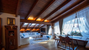 A spacious loft with a high ceiling, wooden beams, a cozy seating area with a sofa and chairs, a dining table, and large windows with sheer curtains. - Elegant Ampezzo-style apartment with breathtaking views of the Dolomites. (Cortina d'Ampezzo)