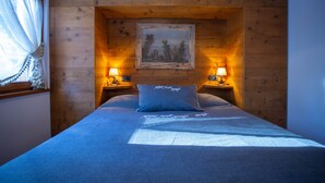 A bedroom with a wooden headboard, a framed picture above the bed, two bedside lamps, and a window with a sheer curtain. - Elegant Ampezzo-style apartment with breathtaking views of the Dolomites. (Cortina d'Ampezzo)